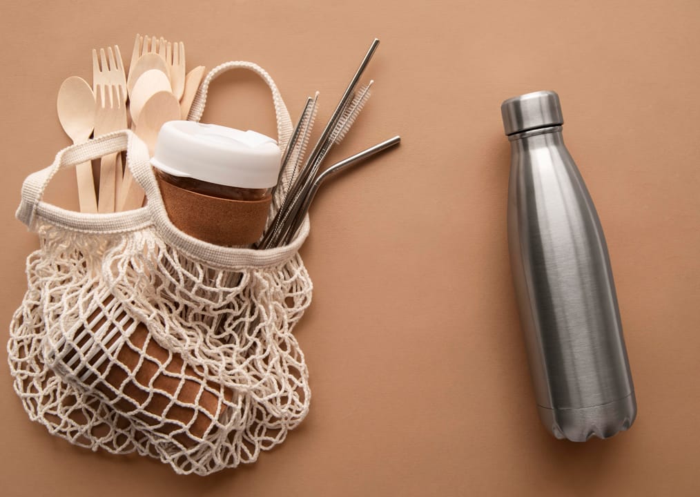 Reusable set – cotton mesh bag, bamboo cutlery and bottle Cotton mesh bag with reusable cup, bamboo cutlery and a stainless-steel bottle on tan background